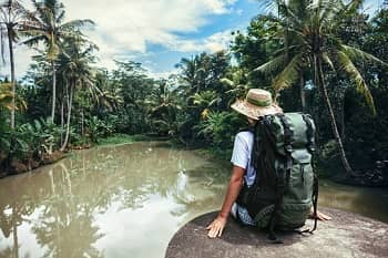 Eine Person mit Reiserucksack und Strohhut sitzt auf einem großen flachen Stein vor einem Gewässer in tropischer Umgebung