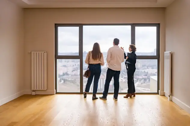 Makler zeigt einem jungen Paar eine Wohnung – Aussicht auf die Stadt aus einem großen Panoramafenster, leeres Zimmer, Hausbesichtigung.