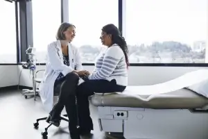 Eine Chefärztin sitzt mit einer Patientin in einem Krankenhauszimmer. Beide sind fröhlich und lachen.