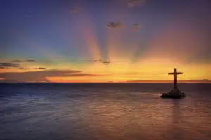 Blick auf das Meer mit einen goldenen Horizont. Im Wasser ragt ein hölzernes Kreuz auf einem kleinen Felsen.